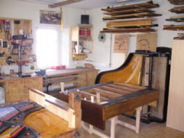 Innenansicht der Werkstatt. Zwei Pianos liegen auf Böcken und warten auf ihre Bearbeitung. An der Wand steht ein Flügel hochkant. An den Wänden sind mit Gehäuseteilen gefüllte Regale angebracht. 