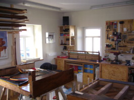Innenansicht der Werkstatt. Zu erkennen sind zwei Pianos, welche auf dem Rücken liegen. Auf der Werkbank steht eine Klaviermechanik. Hinter einer Ständerbohrmaschine ist eine Werkzeugwand zu erkennen. 
